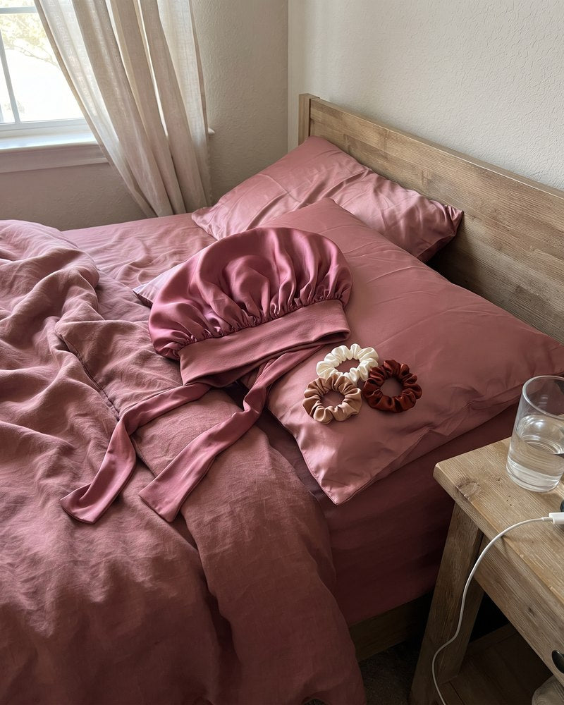 The untied satin bonnet and satin scrunchies on a satin pillowcase in morning light.