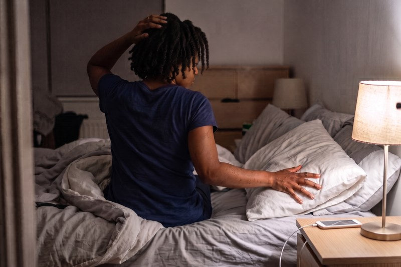 A woman with 4c coils sits up in bed at night, one hand at her bare hair, the other feeling across the pillow beside her.