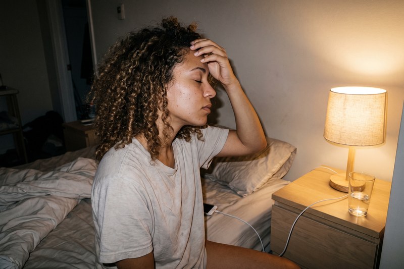 A woman with 3c curls sits on the side of her bed at night, fingertips resting on her forehead.
