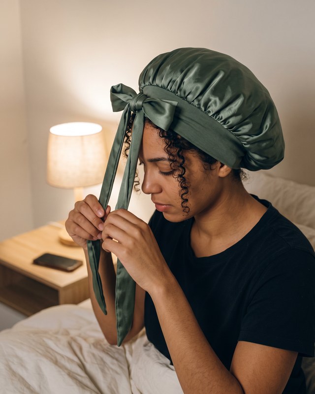 A woman with 3c curls ties the satin bonnet in a bow at the front by lamp light.